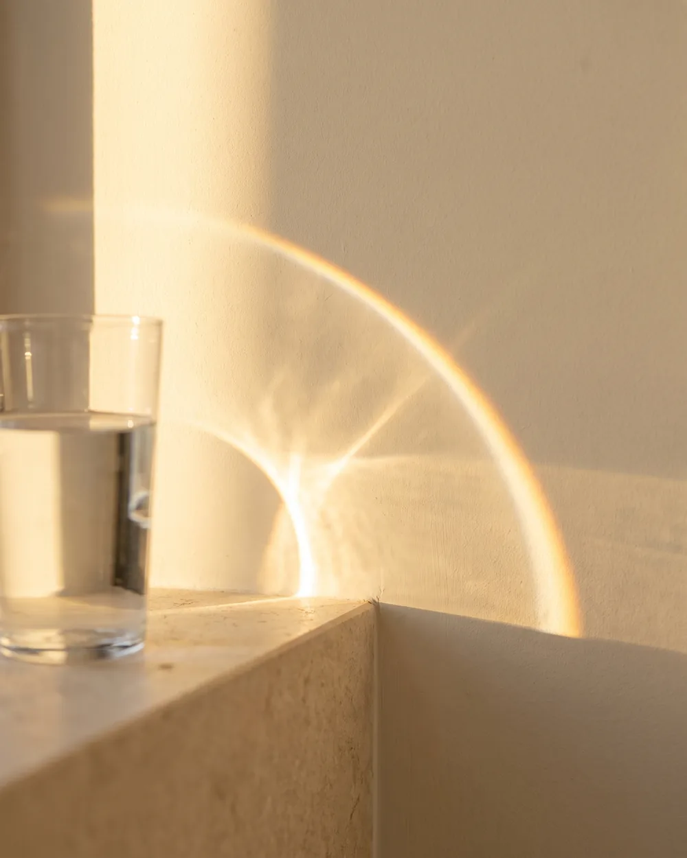 Varmt eftermiddagslys gennem et glas vand der kaster en gylden bue på væggen
